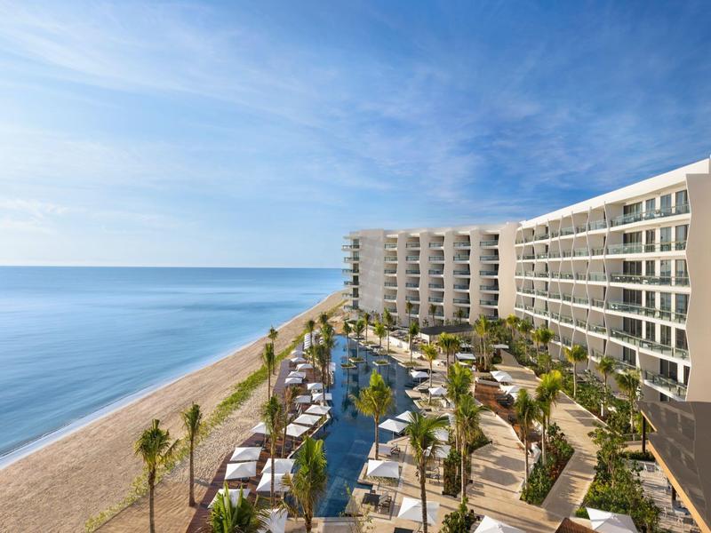 Modernes Strandhotel mit Pool, Palmen und weitläufigem Sandstrand unter klarem Himmel.