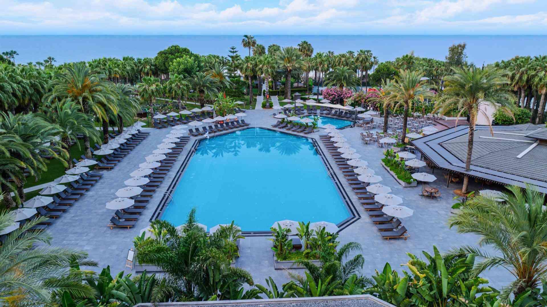 Großer Hotelpool mit Liegestühlen, umgeben von Palmen und Meerblick im Hintergrund.