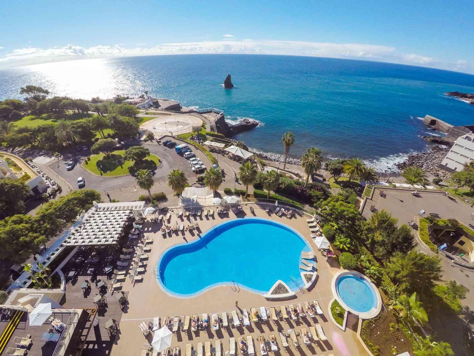Hotelpool mit Liegen, Palmen und Blick auf das weite, blaue Meer unter klarem Himmel.