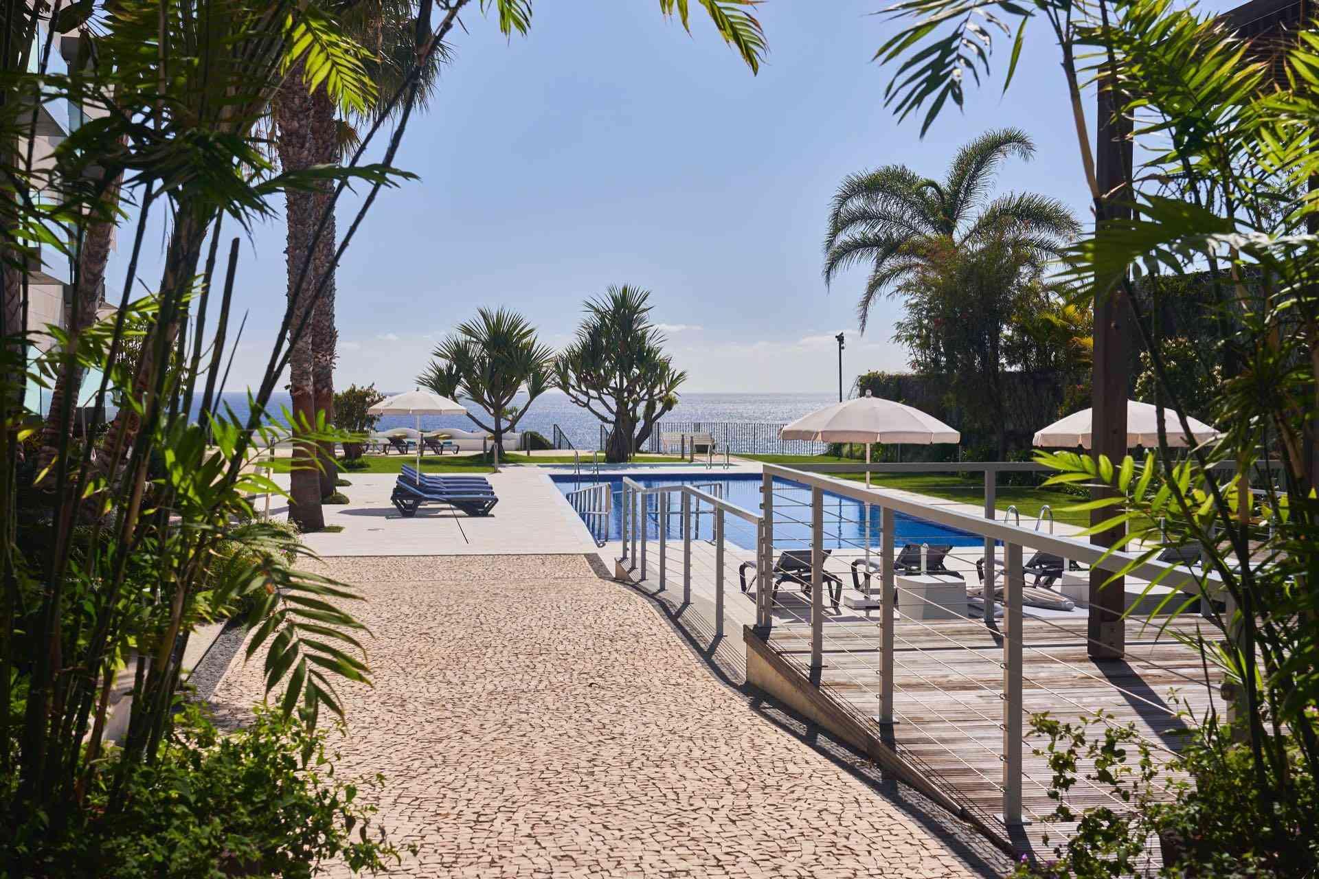 Moderner Hotelpool mit Liegestühlen, Palmen und Aussicht auf das Meer an sonnigem Tag.