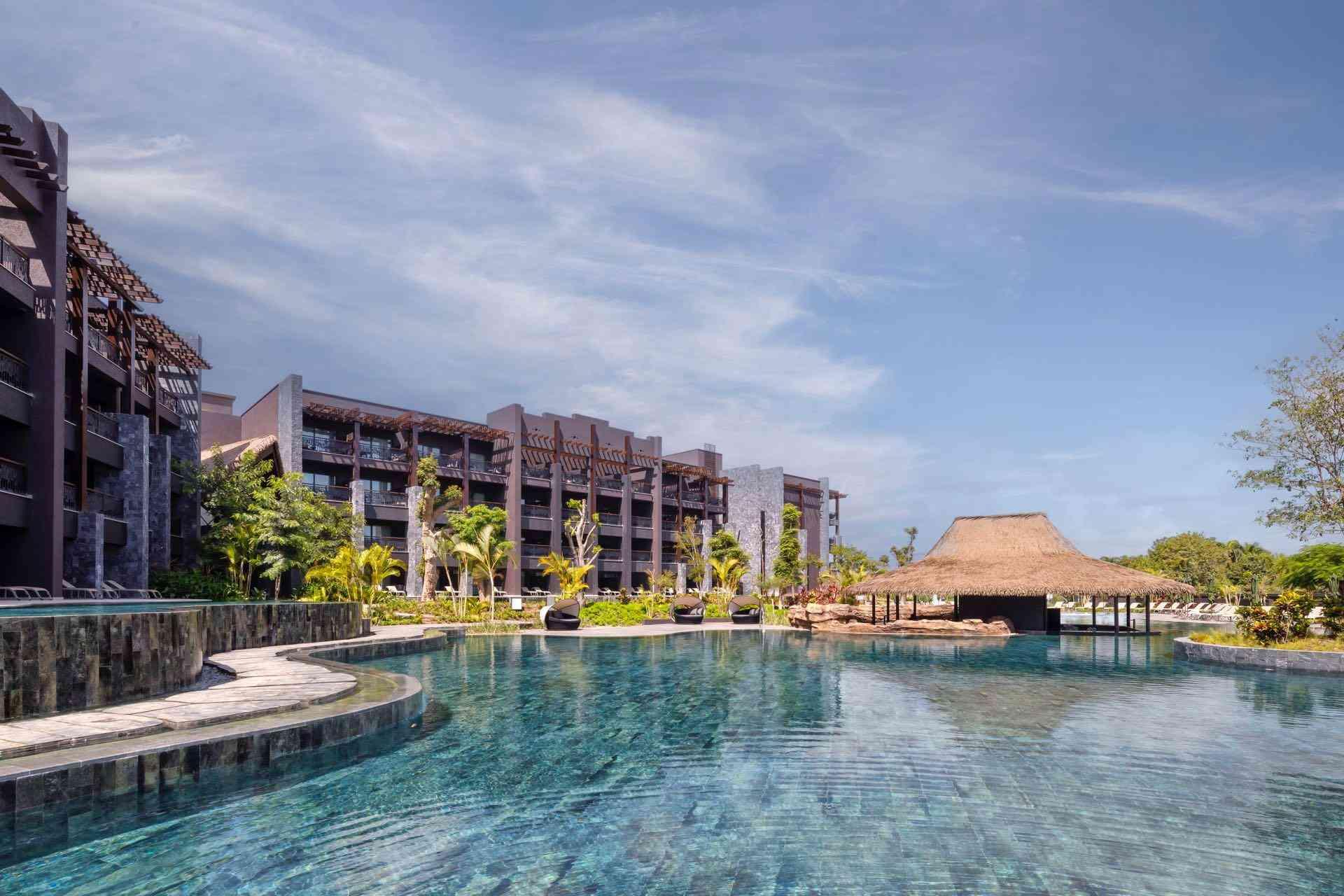 Moderner Hotelkomplex mit großem, klarem Pool und tropischen Palmen unter blauem Himmel.