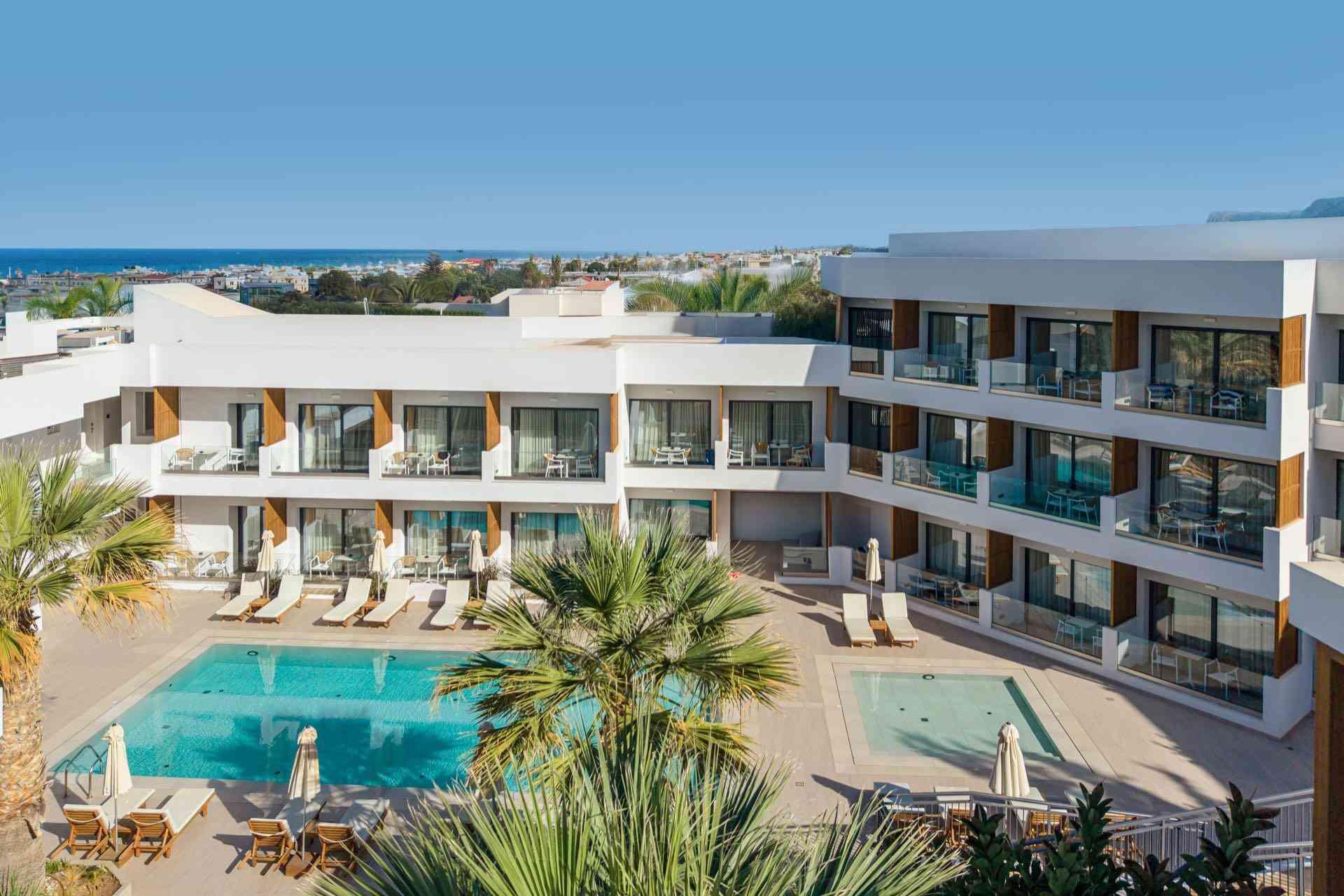 Moderner Hotelkomplex mit Pool, Liegestühlen und Meerblick an klar blauem Himmel.