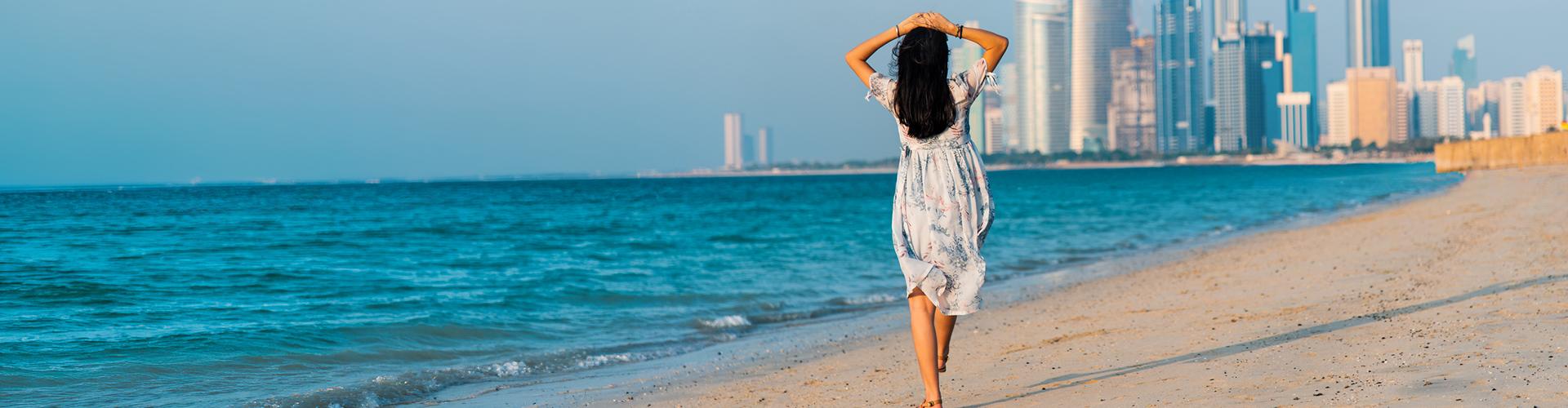 Frau in Sommerkleid spaziert am Sandstrand, blauer Himmel, Meer und Hochhäuser einer Stadt im Hintergrund.