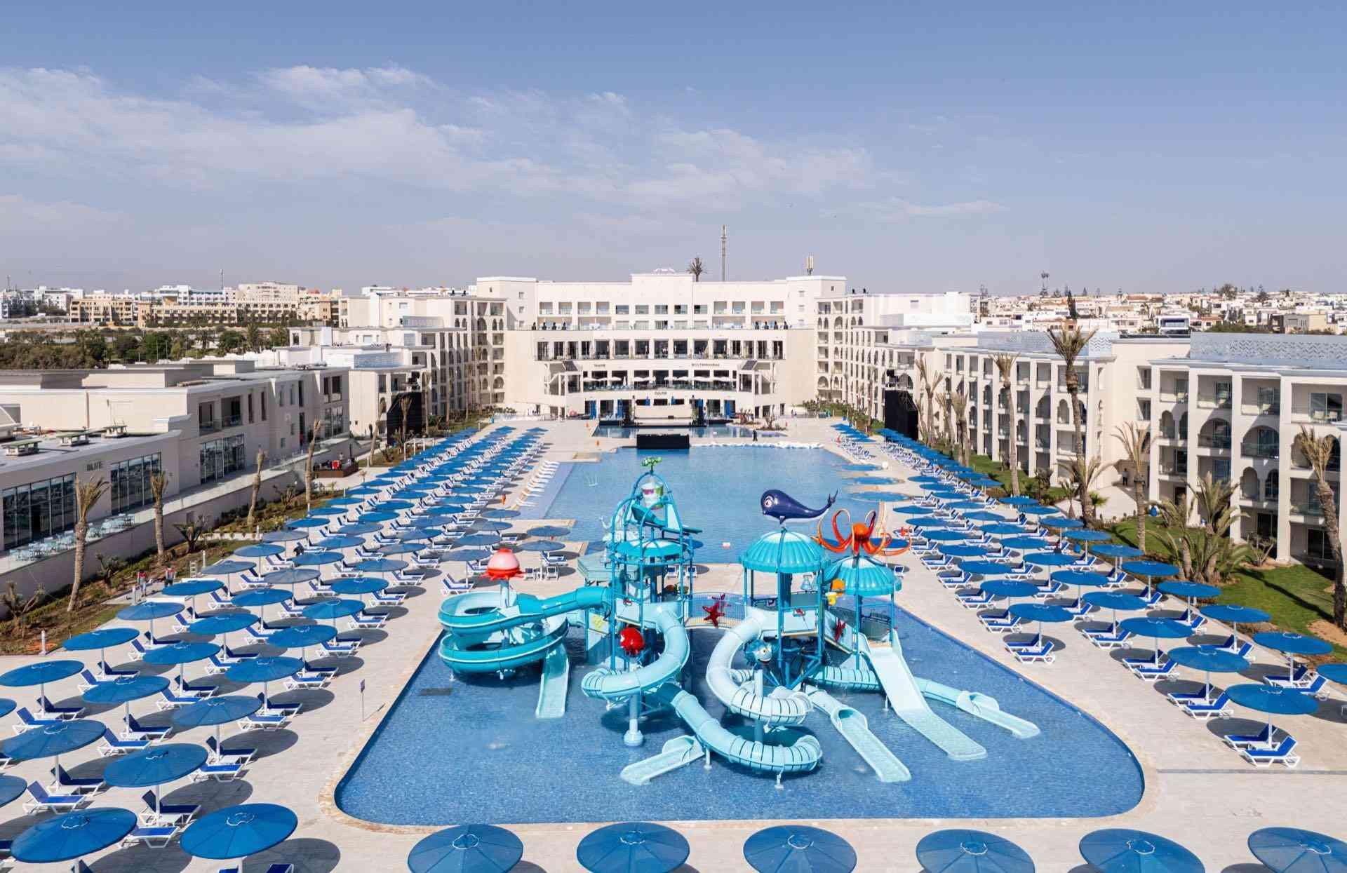 Großes Hotel mit zentralem Pool, Wasserrutschen und zahlreichen blauen Liegestühlen im Außenbereich.