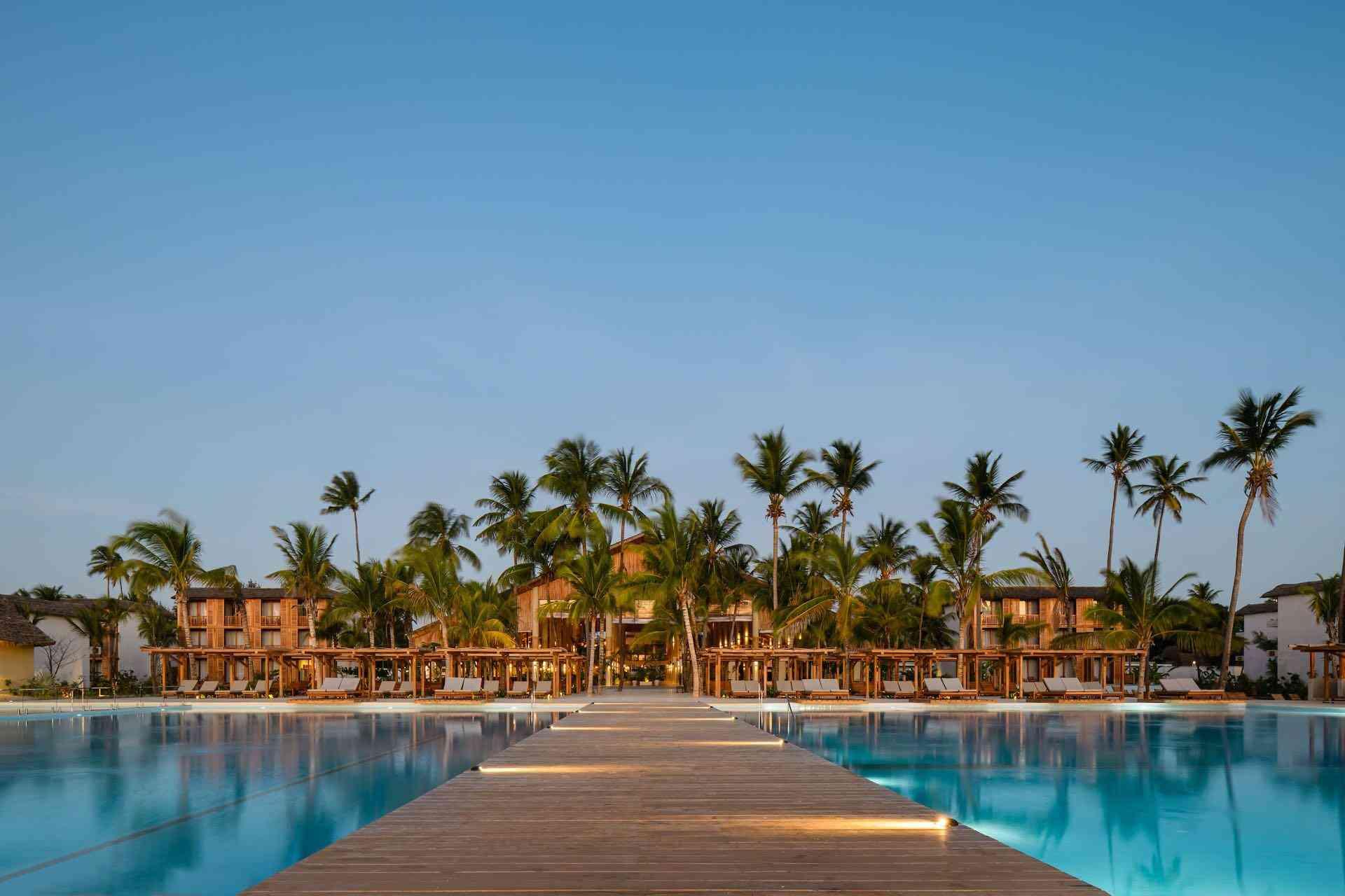 Luxuriöses Hotel mit großem blauem Pool und Holzsteg, umgeben von Palmen, unter klarem Himmel.