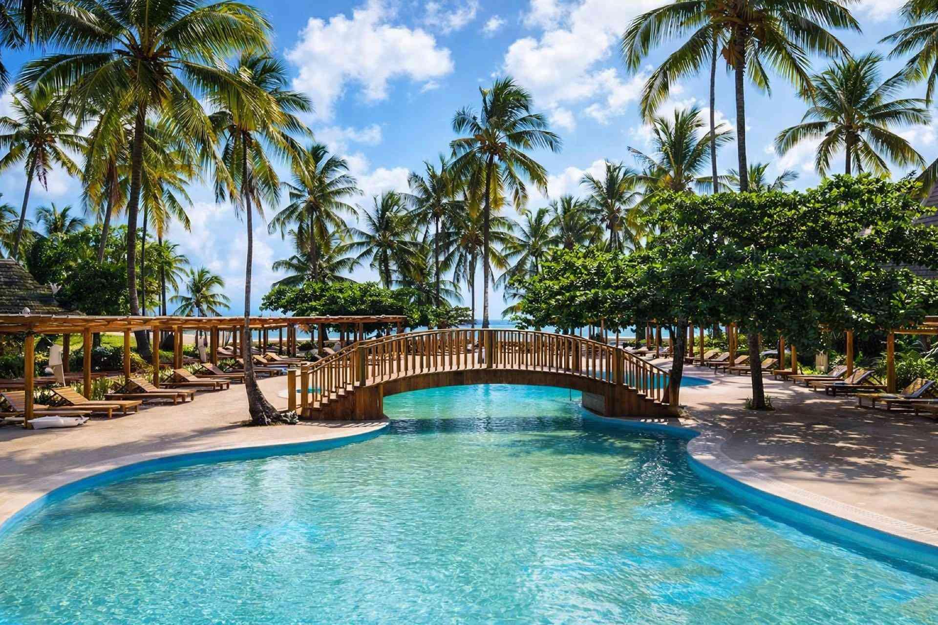 Tropischer Hotelpool mit Holzbrücke, Palmen und Liegestühlen unter blauem Himmel.