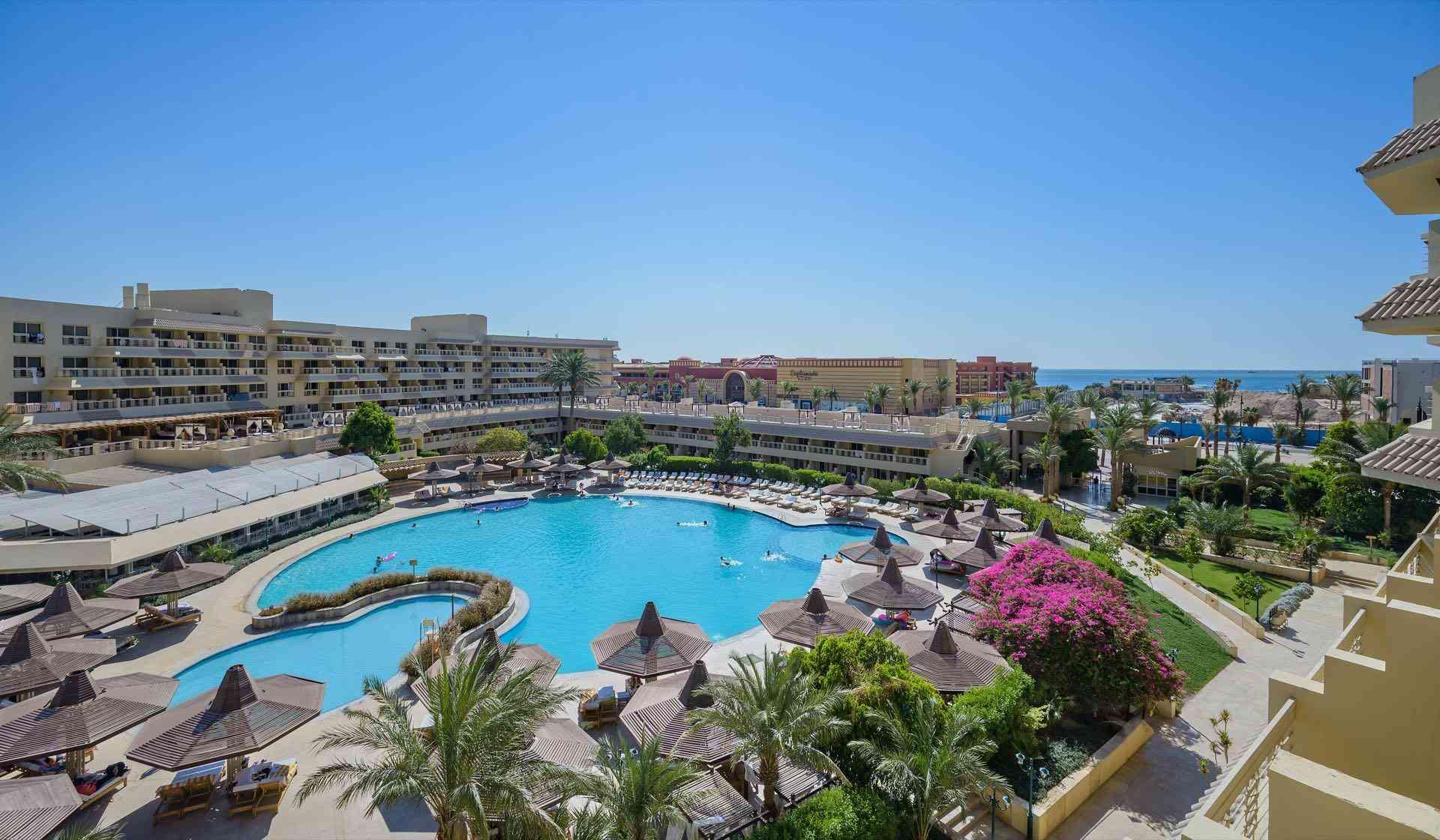 Großer Hotelpool mit Liegen, Palmen und Blick auf Meer unter klarem blauem Himmel.
